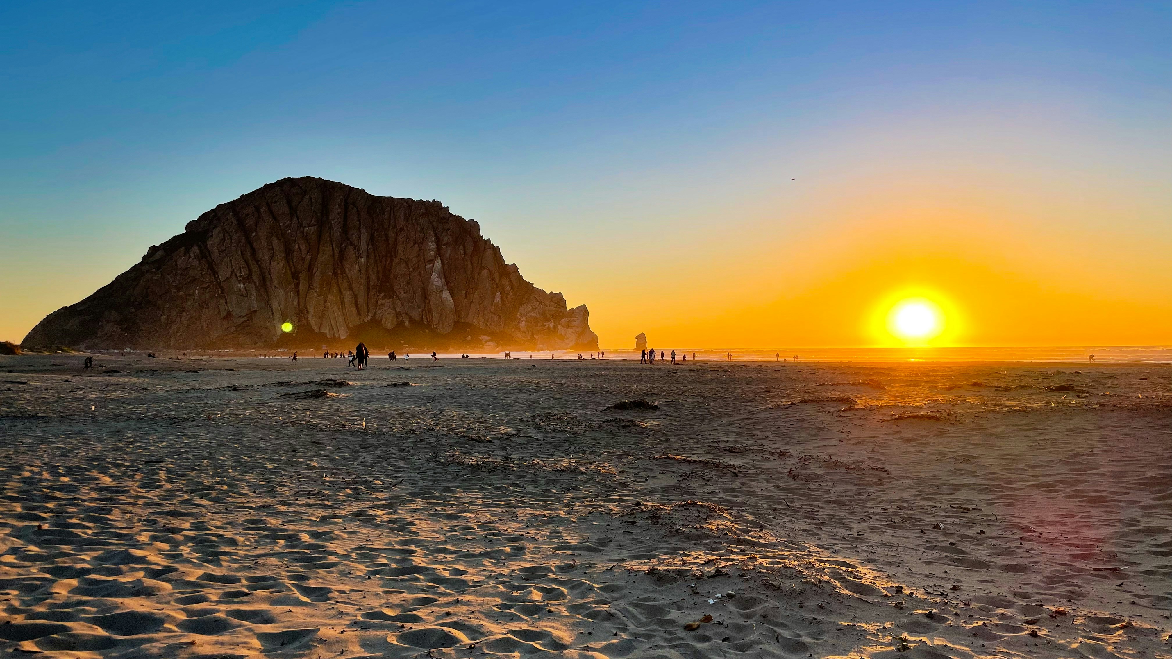 the sun is setting on a beach with a large rock in the background