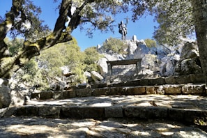 A stone pathway leads through a natural setting with trees on either side, culminating in a statue of a man holding a cross. The scene combines natural elements like trees and rocks with a historical monument, creating a blend of history and nature.