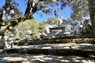 A stone pathway leads through a natural setting with trees on either side, culminating in a statue of a man holding a cross. The scene combines natural elements like trees and rocks with a historical monument, creating a blend of history and nature.