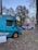 Two food trucks are parked on a grassy area surrounded by trees with sparse leaves. One truck is teal and advertises burgers, while the other is pink and blue, advertising ice cream. An orange traffic cone is positioned nearby, and the sky is clear, indicating late afternoon or early evening.