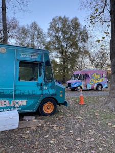 Two food trucks are parked on a grassy area surrounded by trees with sparse leaves. One truck is teal and advertises burgers, while the other is pink and blue, advertising ice cream. An orange traffic cone is positioned nearby, and the sky is clear, indicating late afternoon or early evening.