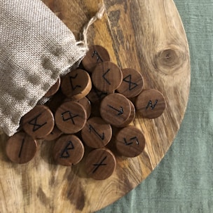 A wooden surface holds several round wooden tokens with ancient runic symbols. The tokens spill out of a small burlap sack placed on the side. The background includes a textured green fabric.