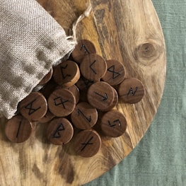 A wooden surface holds several round wooden tokens with ancient runic symbols. The tokens spill out of a small burlap sack placed on the side. The background includes a textured green fabric.
