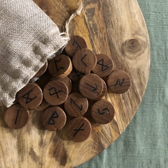 A wooden surface holds several round wooden tokens with ancient runic symbols. The tokens spill out of a small burlap sack placed on the side. The background includes a textured green fabric.