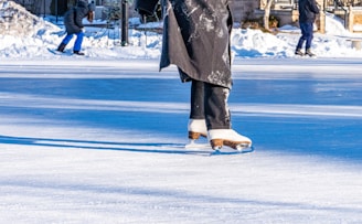 A person wearing white ice skates and dark pants glides gracefully across an outdoor ice rink. The rink is surrounded by snow, with other people in the background also skating. The bright sunlight casts clear shadows on the ice, enhancing the winter atmosphere.