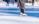 A person wearing white ice skates and dark pants glides gracefully across an outdoor ice rink. The rink is surrounded by snow, with other people in the background also skating. The bright sunlight casts clear shadows on the ice, enhancing the winter atmosphere.