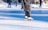 A person wearing white ice skates and dark pants glides gracefully across an outdoor ice rink. The rink is surrounded by snow, with other people in the background also skating. The bright sunlight casts clear shadows on the ice, enhancing the winter atmosphere.