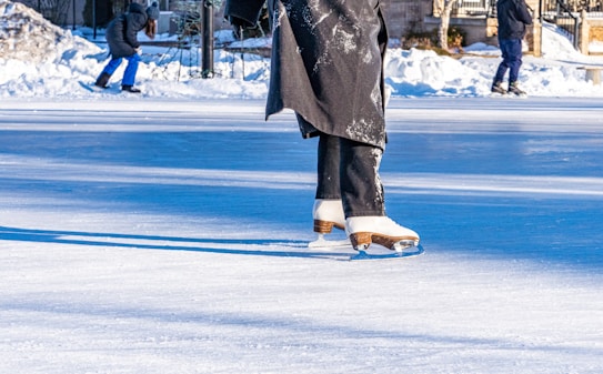 A person wearing white ice skates and dark pants glides gracefully across an outdoor ice rink. The rink is surrounded by snow, with other people in the background also skating. The bright sunlight casts clear shadows on the ice, enhancing the winter atmosphere.