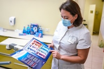 A dental professional wearing a face mask examines a product brochure. The background contains various dental care products including mouthwash and toothpaste packaging placed on a counter. The setting appears to be a dental clinic with a dental chair visible.