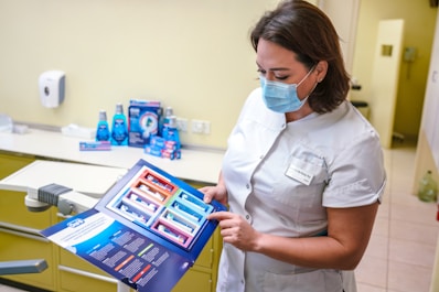 A dental professional wearing a face mask examines a product brochure. The background contains various dental care products including mouthwash and toothpaste packaging placed on a counter. The setting appears to be a dental clinic with a dental chair visible.