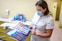 A dental professional wearing a face mask examines a product brochure. The background contains various dental care products including mouthwash and toothpaste packaging placed on a counter. The setting appears to be a dental clinic with a dental chair visible.
