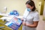 A dental professional wearing a face mask examines a product brochure. The background contains various dental care products including mouthwash and toothpaste packaging placed on a counter. The setting appears to be a dental clinic with a dental chair visible.