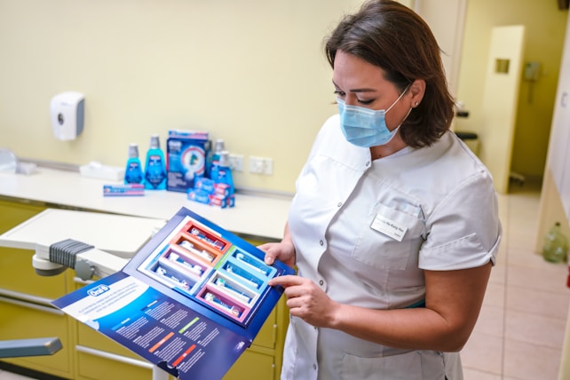 A dental professional wearing a face mask examines a product brochure. The background contains various dental care products including mouthwash and toothpaste packaging placed on a counter. The setting appears to be a dental clinic with a dental chair visible.