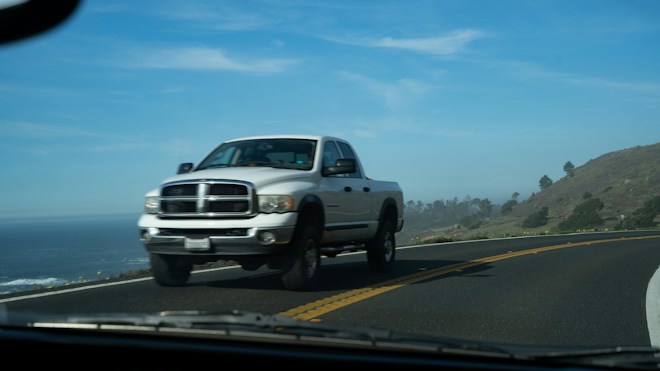 A white pickup truck drives along a winding coastal road surrounded by ocean views and rolling hills. The sky is clear with a few clouds, creating a scenic backdrop.