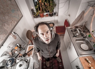 A person stands in a compact kitchen with a slightly cluttered appearance, looking upward towards the camera. The kitchen features a stove with pots and utensils, a sink with dishes, cutting boards with ingredients, and a plant by the window. A map is visible on the wall and there are strings of lights decorating the space.
