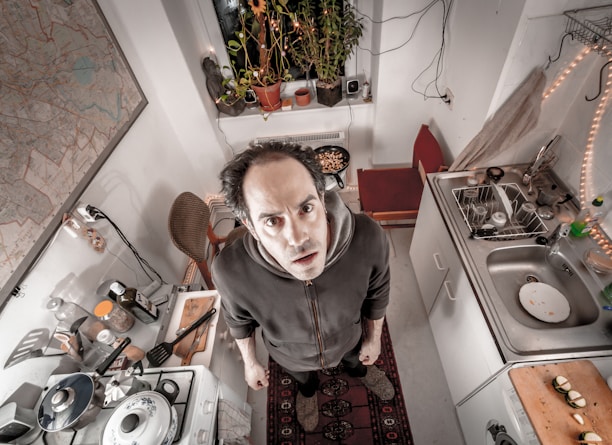 A person stands in a compact kitchen with a slightly cluttered appearance, looking upward towards the camera. The kitchen features a stove with pots and utensils, a sink with dishes, cutting boards with ingredients, and a plant by the window. A map is visible on the wall and there are strings of lights decorating the space.