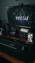 A computer setup featuring a large monitor displaying a dark forest-themed wallpaper with the word 'Hustle' in the center. On the desk, there is a keyboard, a mouse, and a smartphone on a stand displaying various app screens. The room has a modern, tech-focused aesthetic with dim lighting and a speaker on the desk.