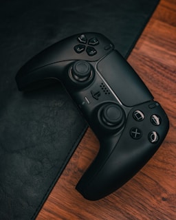 A black video game controller with numerous buttons and joysticks rests on a wooden surface. The controller is slightly tilted, showing its ergonomic design, and is partially surrounded by a dark leather-like material.