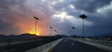 A highway with solar street lights stretches into the distance under a dramatic sky. The sun sets on the left, casting an orange glow amidst dark clouds, creating a striking contrast.