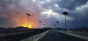 A highway with solar street lights stretches into the distance under a dramatic sky. The sun sets on the left, casting an orange glow amidst dark clouds, creating a striking contrast.
