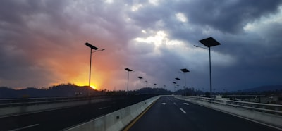 A highway with solar street lights stretches into the distance under a dramatic sky. The sun sets on the left, casting an orange glow amidst dark clouds, creating a striking contrast.