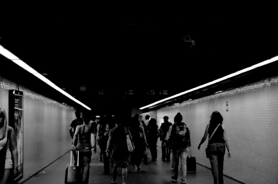 A group of people walking through a dimly-lit underground corridor with luggage. The corridor is tiled and narrow, with fluorescent lights running along the ceiling. The atmosphere feels busy yet calm, as individuals move through the space.