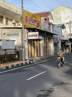 A street view showing a person riding a motorcycle along an urban road. On the side of the street, there are several shops with signs, including one labeled 'Pelangi Baby Shop'. The buildings are relatively old with faded paint and some show signs of wear. Electrical wires crisscross overhead, casting shadows on the street.