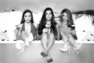 Three women are sitting in a bathtub, each holding a cocktail. They have relaxed expressions and are dressed stylishly, with their legs stretched out over the edge. The background features a tiled wall and scattered rose petals, adding a touch of elegance.