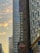 A vertical marquee sign for the Nederlander Theatre against a backdrop of city skyscrapers. The sign is ornately designed with colorful patterns and lettering. To the left, the sky displays a soft evening glow with few clouds, and modern high-rise buildings fill the scene.