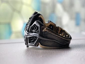 A close-up of an ornate metal ring placed on a smooth surface. The ring features intricate designs, combining silver and dark metallic tones, with a textured and polished finish. The background is softly out of focus, displaying a mosaic pattern of blue and green hues.
