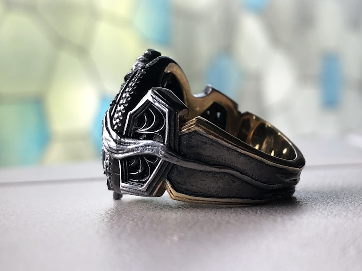A close-up of an ornate metal ring placed on a smooth surface. The ring features intricate designs, combining silver and dark metallic tones, with a textured and polished finish. The background is softly out of focus, displaying a mosaic pattern of blue and green hues.