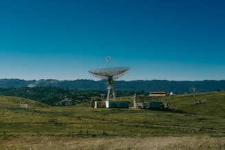 A large satellite dish stands in an open field surrounded by rolling hills and dense forest in the background. Several small buildings and structures are positioned nearby, connected by wires. The sky is clear with a deep blue hue.