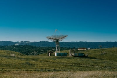 A large satellite dish stands in an open field surrounded by rolling hills and dense forest in the background. Several small buildings and structures are positioned nearby, connected by wires. The sky is clear with a deep blue hue.