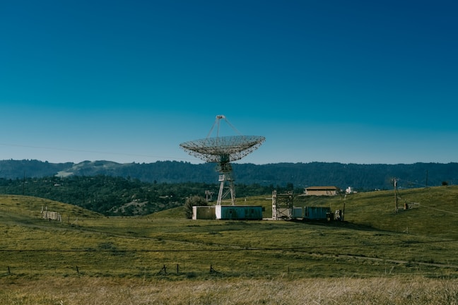 A large satellite dish stands in an open field surrounded by rolling hills and dense forest in the background. Several small buildings and structures are positioned nearby, connected by wires. The sky is clear with a deep blue hue.