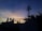 A telecommunications tower stands silhouetted against a vibrant sunset sky. The sky displays a gradient of deep blues transitioning into warm oranges and yellows near the horizon. In the foreground, various plants and the outline of a building with signage are visible.