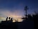 A telecommunications tower stands silhouetted against a vibrant sunset sky. The sky displays a gradient of deep blues transitioning into warm oranges and yellows near the horizon. In the foreground, various plants and the outline of a building with signage are visible.