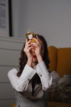 A person with long, curly hair is holding up a bottle of perfume in front of their face. They are wearing a white shirt with black buttons and a black tie, along with a textured vest. There is a yellow chair and a plant in the background.
