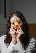 A person with curly hair holds a bottle of Chanel N°5 perfume in front of their face. Their nails are painted red, and they are wearing a white shirt with a black and white vest. The background is softly blurred.