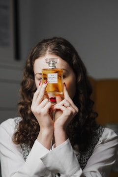 A person with curly hair holds a bottle of Chanel N°5 perfume in front of their face. Their nails are painted red, and they are wearing a white shirt with a black and white vest. The background is softly blurred.