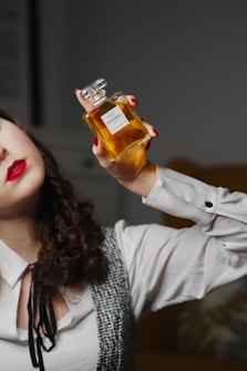 A person with curly hair and red lipstick holds a bottle of Chanel No. 5 perfume in a sophisticated pose. They wear a white shirt with black trim and a patterned vest.