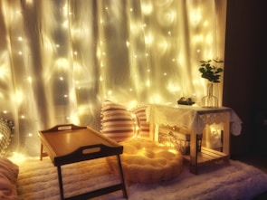 A cozy and warm setting featuring soft yellow fairy lights draped over a white curtain. There is a wooden tray table on a fluffy carpet, a striped throw pillow, and a round, knitted pouf. A small wooden table is adorned with a lace tablecloth and holds a vase with green leaves and decorative items. The ambiance suggests a relaxing and inviting atmosphere.