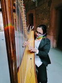 A person is playing a harp, sitting indoors against a stone wall backdrop. They seem focused on the instrument, and there's a rainbow refraction visible on both the harp and the person.