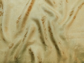 A close-up view of draped, textured fabric with a shimmering, light yellow-gold hue. The surface features subtle floral patterns in a darker shade, creating an elegant and soft appearance.