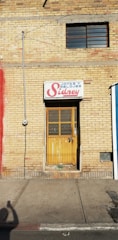 A yellow brick wall with a narrow door and a sign above it that reads 'Joyas y Relojes Sidney'. The door is wooden with a metal grill and paint is chipped at the bottom. There is a single window with metal bars above the door. A utility meter and wiring are seen on the left side of the wall, with a shadow of a person taking a photo cast on the sidewalk.