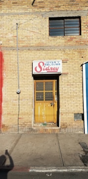 A yellow brick wall with a narrow door and a sign above it that reads 'Joyas y Relojes Sidney'. The door is wooden with a metal grill and paint is chipped at the bottom. There is a single window with metal bars above the door. A utility meter and wiring are seen on the left side of the wall, with a shadow of a person taking a photo cast on the sidewalk.