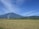 A vast field of ripe golden crops stretches out under a clear blue sky. In the background, a large mountain dominates the horizon, partially shrouded in haze. A plume of white smoke rises near a cluster of small structures at the base of the mountain. Several poles dot the landscape, possibly for electrical wires.
