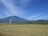 A vast field of ripe golden crops stretches out under a clear blue sky. In the background, a large mountain dominates the horizon, partially shrouded in haze. A plume of white smoke rises near a cluster of small structures at the base of the mountain. Several poles dot the landscape, possibly for electrical wires.