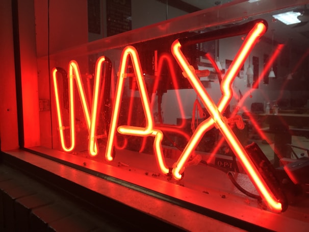 Bright red neon sign displaying the word 'WAX', set against a reflective glass surface, with a glimpse into a dimly lit room behind the sign.