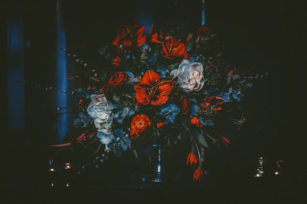A dark, moody floral arrangement featuring a mix of red, white, and dark green flowers. The bouquet is densely packed and placed in a vase with two small candles nearby, creating a dramatic, almost gothic ambiance.