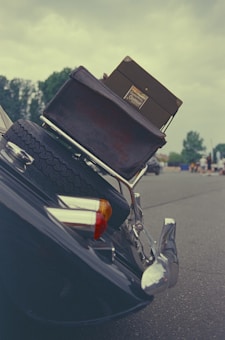 A vintage car's rear is shown with a tire affixed to the back. An antique suitcase is strapped on top, and the background features a parking lot or open space with some trees and distant figures.